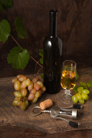 The concept of a delicious alcoholic drink, wine and grapes. Wine bottle, cork, corkscrew, glass of white wine and bunches of pink and green grapes on dark rustic wooden background with grapes leaves.の写真素材
