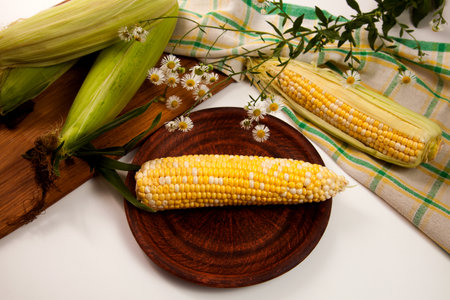 Clay plate with ear of ripe sweet corn and green towel on vintage white wooden background. Cobs with...