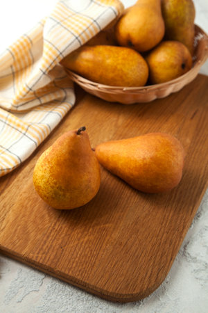 Wooden cutting board with ripe juicy pear fruits, wicker basket with pears and yellow kitchen towel on white wooden background.の写真素材