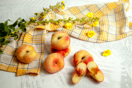 Halves and several whole ripe chinese flat peach fruits or saturn peaches on vintage white wooden background with yellow kitchen towel and yellow flowers.の写真素材