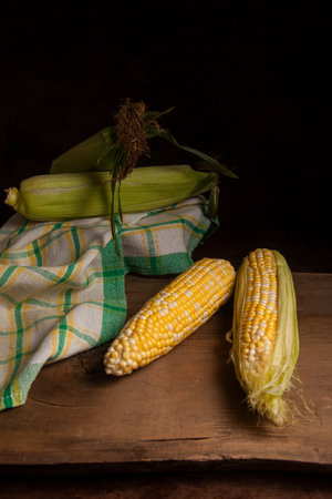 Two ears of ripe sweet corn with green leaves and green towel on vintage wooden background. Cobs with white and yellow grains.の写真素材