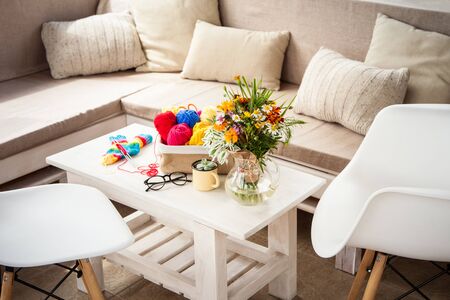 Bright colored yarn for knitting in a basket. A bouquet of wild flowers. Glasses, chairs. White...