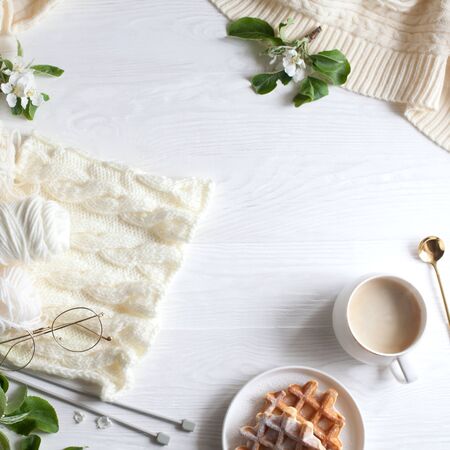 Knitting, knitting needles, yarn, coffee and waffles stand on a white wooden background. Surrounded by branches of a blossoming apple tree. Flatlayの写真素材