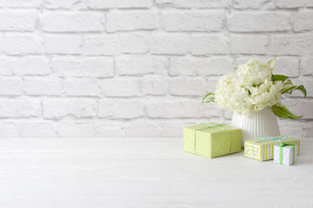 Little green gifts and a bouquet of white hydrangea in a vase. brick wall and white wooden table.の写真素材