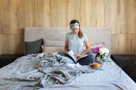 Woman sitting in bed reading a book and having breakfast.の写真素材