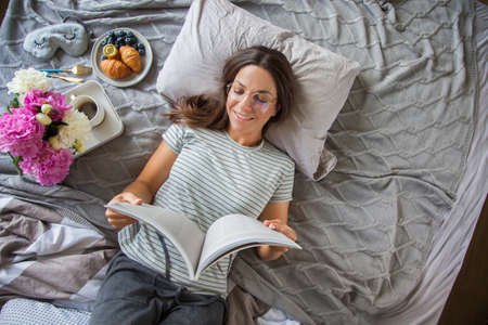 attractive woman lies on bed in the morning, drinking coffee, reading book, casual style, grey dress, feeling comfortable at home, having rest, smilingの写真素材