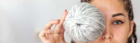 A beautiful young girl knitter smiles and holds skeins of light gray and white yarn in her hands. Woman with dreadlocks.の写真素材