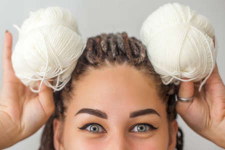 A beautiful young girl knitter smiles and holds skeins of white yarn in her hands, applying them to her head like the ears of a hare or a mouse. Woman with dreadlocks.の写真素材