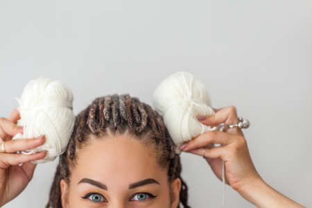 A beautiful young girl knitter smiles and holds skeins of white yarn in her hands, applying them to her head like the ears of a hare or a mouse. Woman with dreadlocks.の写真素材