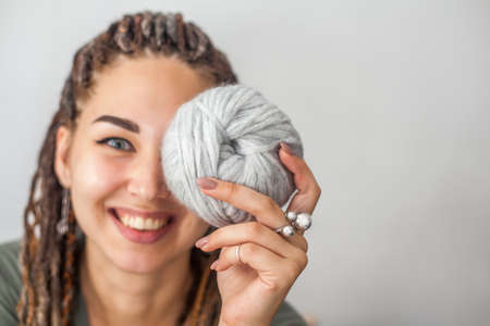 A beautiful young girl knitter smiles and holds skeins of light gray and white yarn in her hands. Woman with dreadlocks.の写真素材