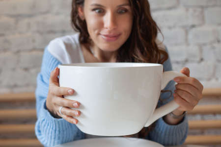 Young woman in blue sweater is feeling headache, with head in hand and holding big cup hot drink.の写真素材