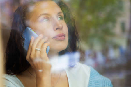 View through window of young woman talking on phone and working on computer with cup of hot coffee.の写真素材