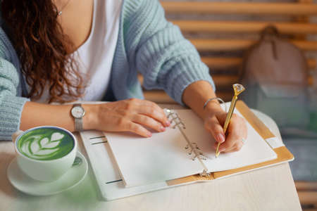 Well groomed woman hand holding gold pen and writing notes with gold pen in notebook. cup of green spirulina latteの写真素材