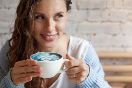 Happy young woman in knitted warm sweater holding with hands a cup of healthy hot blue coffee latte