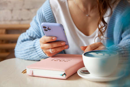 Young woman in blue warm sweater is reading a text message from mobile phone while drinking coffeeの写真素材