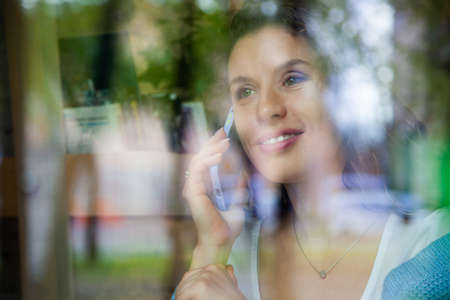 View through window of young woman talking on phone and working on computer with cup of hot coffee.