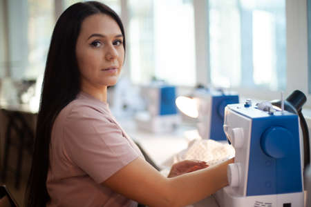 Beautiful woman tailor with long dark hair completing work with sewing machine at workplaceの写真素材