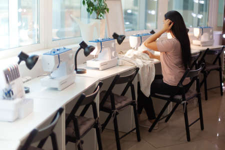 Beautiful woman tailor with long dark hair completing work with sewing machine at workplaceの写真素材