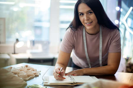 Beautiful young woman designer is making notes of clothing measurements on notebook in sewing studio