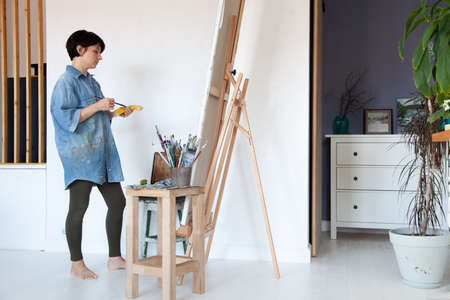 Young woman artist holding paintbrush and painting with oil paint on framed cotton canvas on easelの写真素材