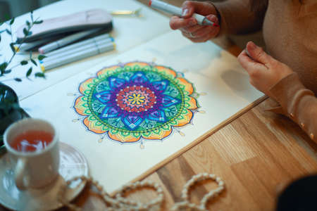 Young woman colouring mandala with markers and white rosary on table with cup of coffee at home