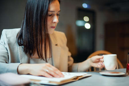 Beautiful young woman in suit writing her business plans while drinking coffee on break timeの写真素材