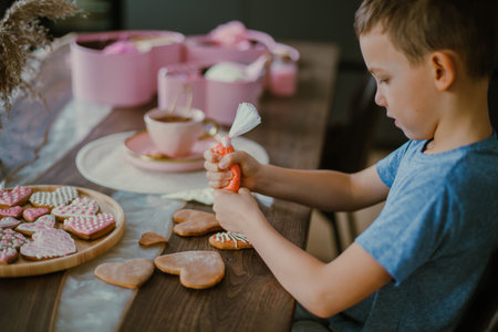 Little cute boy decorates gingerbread heart shaped with pink sugar icing. Preparation for Valentines...