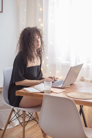 Curly brunette freelancer woman working remotely use laptop at cozy home scandinavian interiorの写真素材