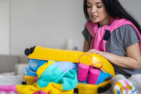 Happy Asian woman trying to closing yellow suitcase full of different clothes accessories and shoesの写真素材