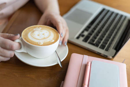 Closeup business woman hands holding cappuccino cup with art picture working at cafe use laptop