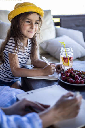 Focused cute little girl in summer yellow hat drawing writing imagination idea on breakfast tableの写真素材
