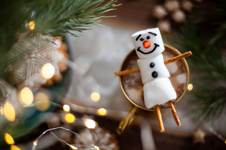 A marshmallow snowman decorated with icing. Gold mug with cocoa and Christmas decor. Gingerbread in...