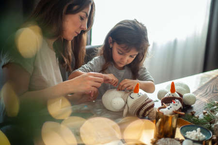 Little cute girl and young beautiful woman make knitted snowmen. Mom and daughter are sewing on buttons.の写真素材