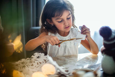 Little cute girl cuts snowflakes from white paper. Gingerbread and cocoa with marshmallows. The concept of preparation for the New Year and Christmas.の写真素材
