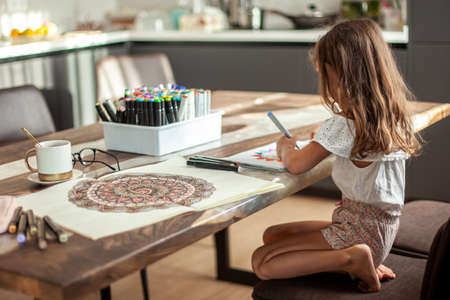 A little cute girl draws a circular mandala pattern in the album with art markers.の写真素材