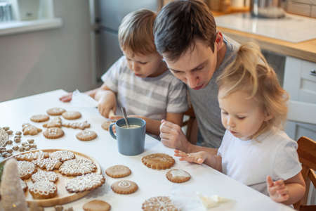 Dad and children decorate Christmas gingerbread at home. A boy and a girl paint with cornets with sugar icing on cookies. New Years decor, branches of a Christmas tree.の写真素材