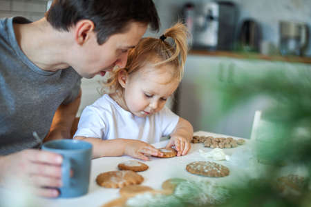 Dad and daughter decorate Christmas gingerbread at home. A boy and a girl paint with cornets with sugar icing on cookies. New Years decor, branches of a Christmas tree.の写真素材