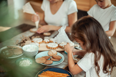 Mom and children decorate Christmas gingerbread at home. A boy and a girl paint with cornets with sugar icing on cookies. New Years decor, branches of a Christmas tree.の写真素材