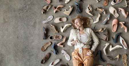 Top view stylish blonde female posing choosing shoes sitting on floor. Top view many female shoes on gray floor fashion stylish footgear choosingの写真素材