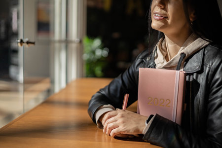 Inspired young woman with a smile looking through window and holding coral colored diary 2022