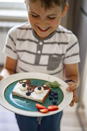 Laughing male kid cooking breakfast decorating fresh sandwich with berries and fruits at kitchenの写真素材
