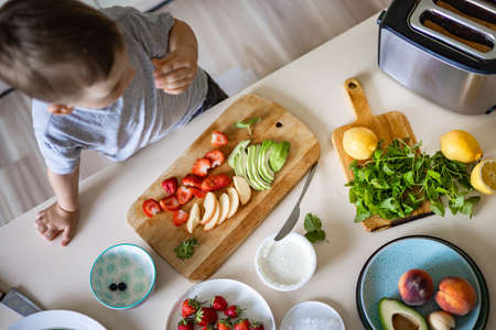 Cheerful boy cooking healthy breakfast at kitchen with avocado, cottage cheese, fruit and berriesの写真素材