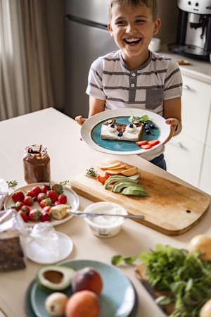 Laughing male kid cooking breakfast decorating fresh sandwich with berries and fruits at kitchenの写真素材