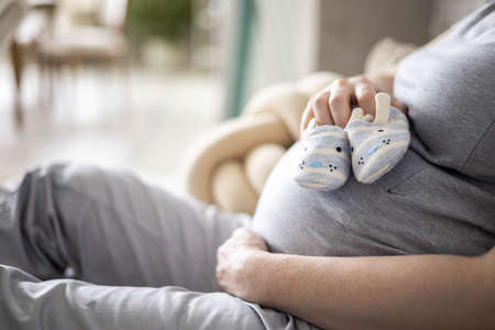 Closeup pregnant female holding cute childish booties on huge tummy relaxing at home awaiting babyの写真素材