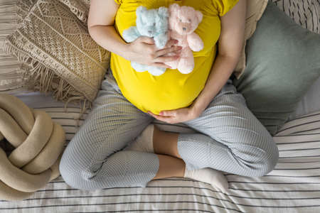 Top view pregnant young woman holding yellow with two cute bear toys on belly sitting on bedの写真素材