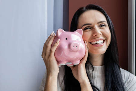 Closeup face laughing young brunette woman holding pink piggy bank rejoicing having positive emotionの写真素材