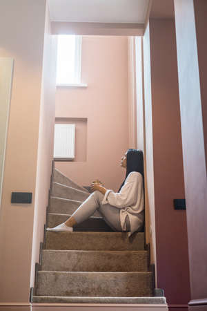 Pensive woman sitting on stairs at home cottage with hot beverage dreaming looking into windowの写真素材