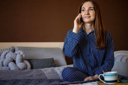 Happy young domestic woman in pajamas sitting on bed talking use smartphone smiling enjoying morning