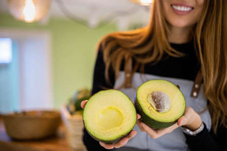 Portrait of smiling young woman vendor in apron holding two halves of fresh ripe green avocadoの写真素材