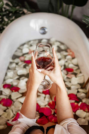 Closeup sexy woman in black lingerie hands holding goblet red wine lying in retro bath roses petals